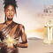 A woman in a shimmering bronze outfit stands confidently in a desert landscape during sunset, holding a feather-like accent. Beside her is a bottle of the "Alien Goddess" perfume.
