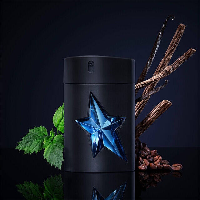 A sleek black fragrance bottle featuring a blue star emblem is positioned against a dark background, accompanied by sticks of wood and scattered coffee beans.