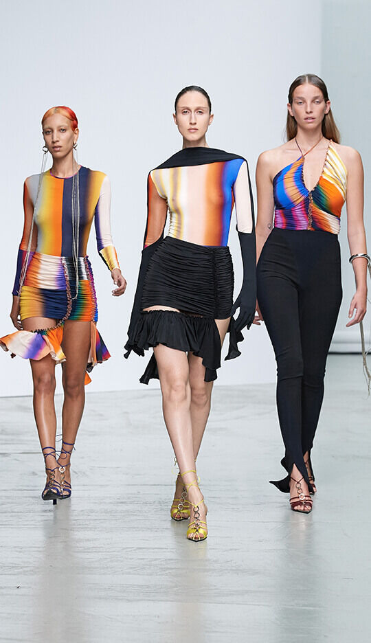 Three models walk down a runway showcasing vibrant, colorful outfits. The first model wears a form-fitting dress with a flared hem, the second has a long-sleeved abstract-pattern top paired with a mini skirt, and the third sports a sleek black ensemble with a halter neckline.