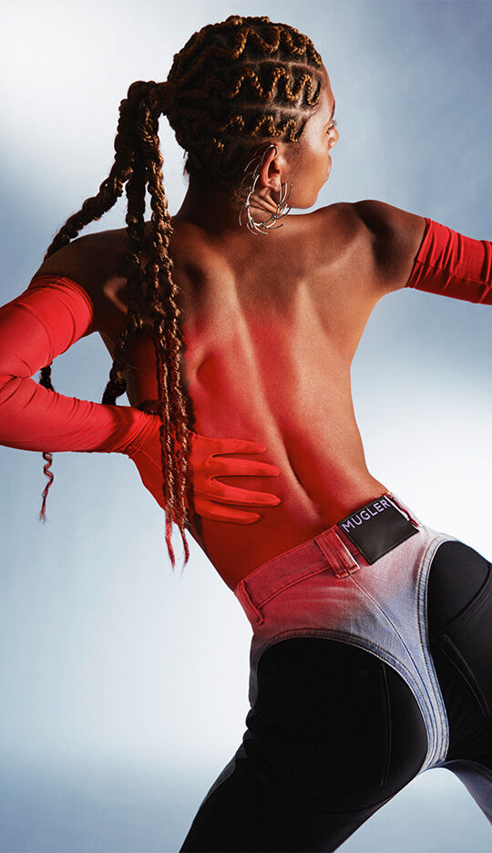 A model poses with her back to the camera, showcasing intricate braided hair and wearing a bold, red long-sleeve top paired with stylish denim pants. The background features soft lighting that enhances the vibrant colors of the outfit.