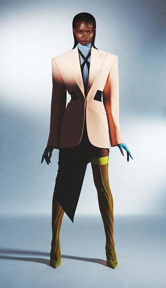 A model stands confidently in an avant-garde outfit featuring an oversized beige blazer, a blue collared shirt, and asymmetrical skirt. The ensemble is completed with colorful thigh-high stockings and coordinating gloves, set against a minimalist background.