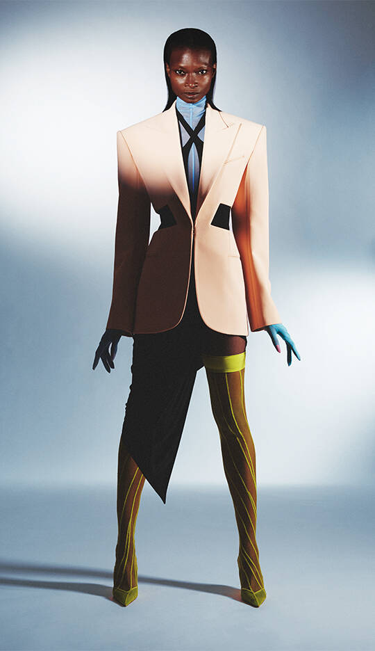 A model stands confidently in an avant-garde outfit featuring an oversized beige blazer, a blue collared shirt, and asymmetrical skirt. The ensemble is completed with colorful thigh-high stockings and coordinating gloves, set against a minimalist background.
