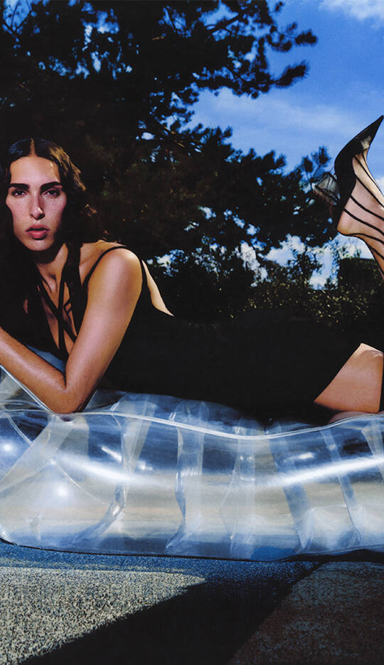 A woman in a black dress lounges on a clear inflatable surface, striking a relaxed pose against a backdrop of trees and a blue sky. She wears high heels and has long, wavy hair.