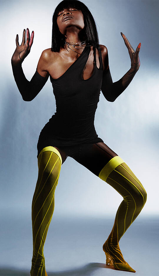 A model poses dramatically against a muted background, wearing a sleek black dress that features one long sleeve and a cut-out. They sport bright yellow thigh-high stockings and striking yellow heels, creating a bold contrast in the styling.