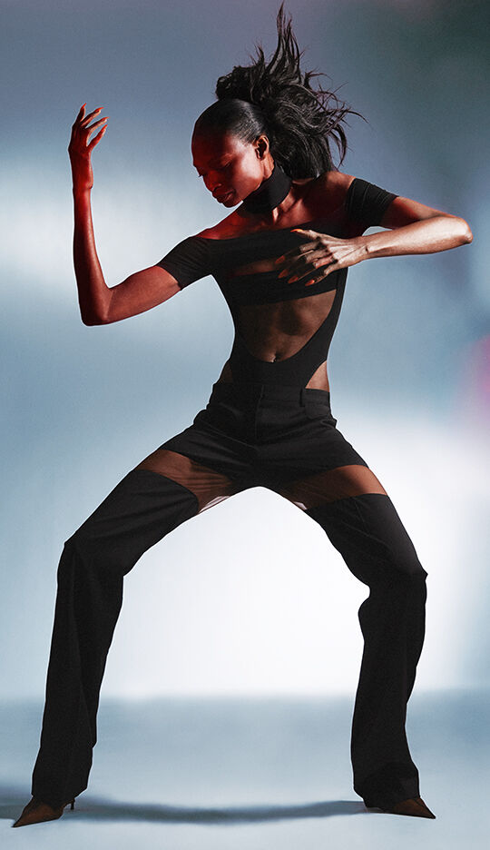 A dancer with a striking pose wears a black outfit featuring sheer panels, set against a softly blurred background. The lighting highlights the fluidity of movement and the contours of the garment.
