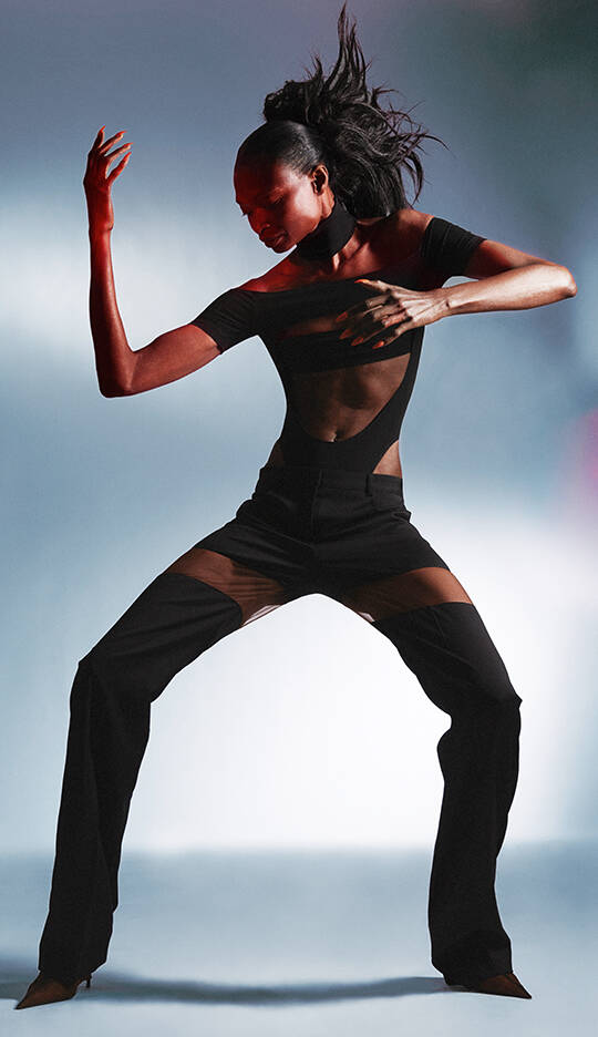 A dancer with a striking pose wears a black outfit featuring sheer panels, set against a softly blurred background. The lighting highlights the fluidity of movement and the contours of the garment.