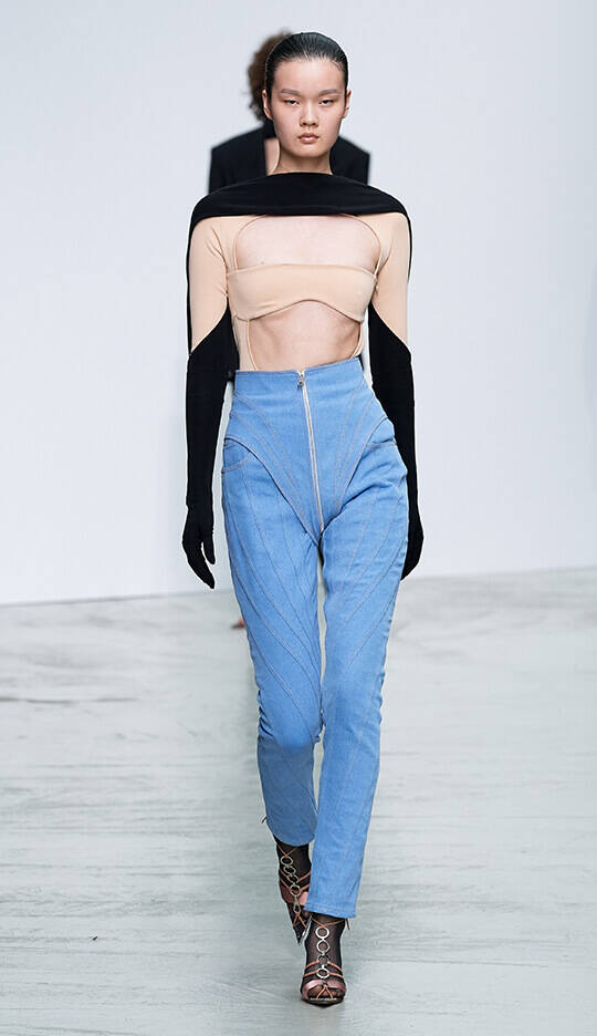 A model walks down the runway, wearing a stylish outfit consisting of a cropped, skin-toned top with black long sleeves and high-waisted, light blue pants featuring a zipper detail.
