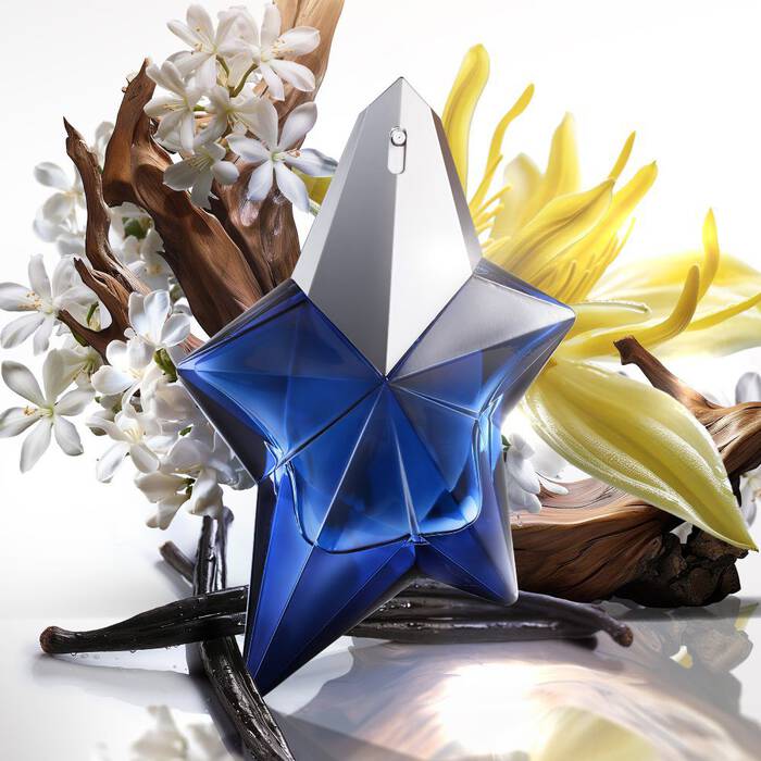 A faceted, star-shaped blue perfume bottle with a silver cap, surrounded by white jasmine flowers, a large yellow orchid, gnarled wood, and dark vanilla bean pods, set against a white background.