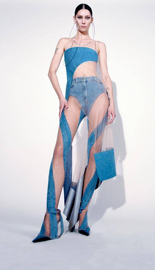 A model poses in a striking blue outfit made of denim and transparent materials, featuring cut-out designs and a long, flowing silhouette.