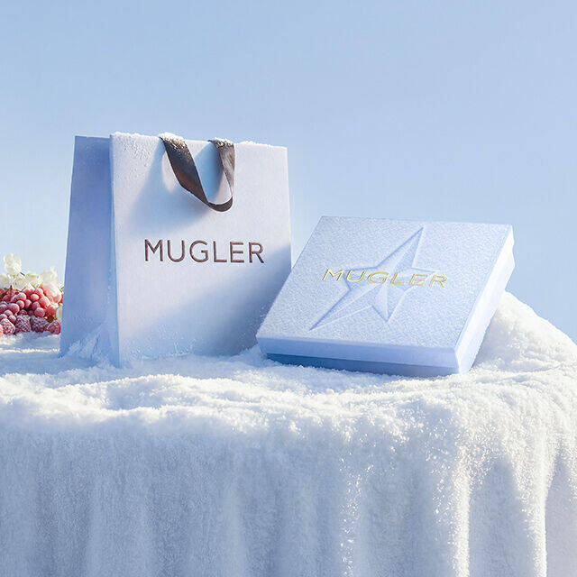 A light blue Mugler shopping bag with dark handles stands next to a flat, square light blue Mugler gift set in the snow. text reads "complimentary shipping. become an insider today to enjoy complimentary shipping every day"