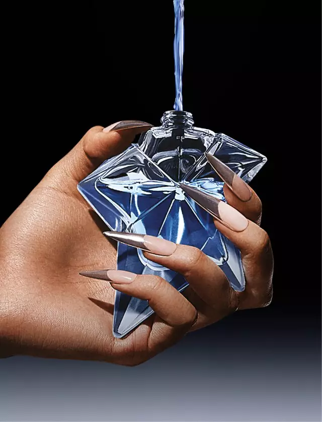 A hand holding a uniquely shaped, blue perfume bottle with a glossy finish against a dark background.