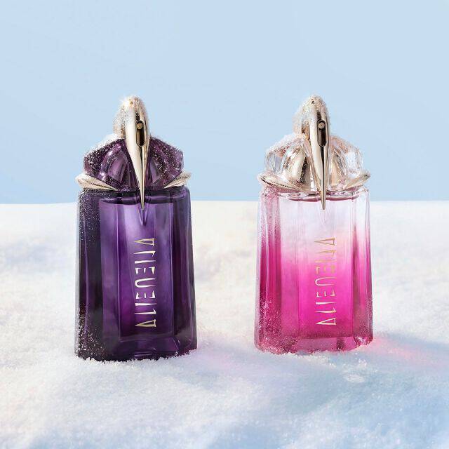 Two Mugler Alien perfume bottles, one classic deep purple (Alien eau de parfum) and one vibrant pink (Alien pulp), standing next to each other on a snowy banquet table. text reads "gifts under $150"