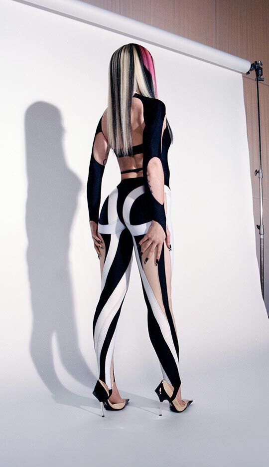 A person facing away from the camera, wearing a fitted black and white outfit with bold patterns, and high-heeled shoes, standing on a photography set with a plain background.