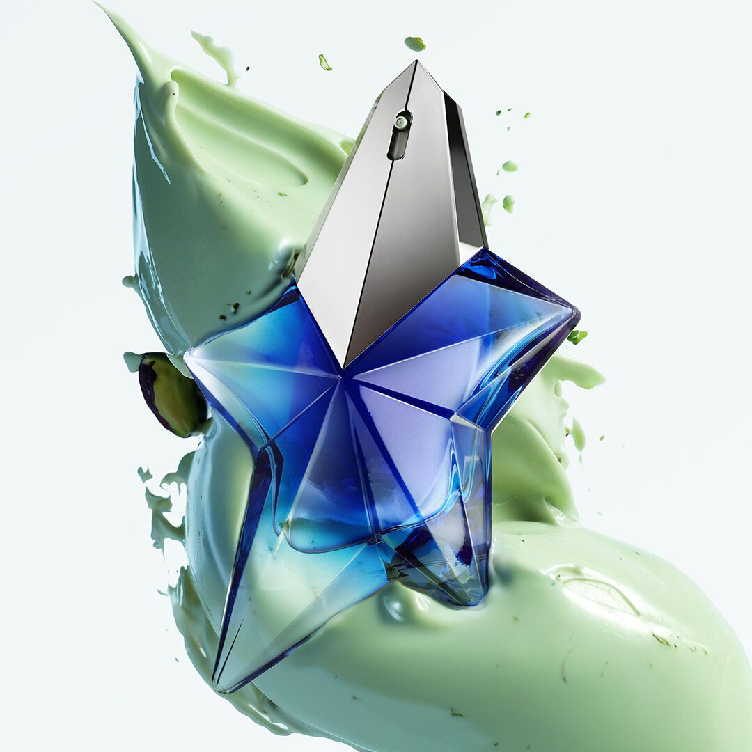 A faceted, star-shaped blue perfume bottle with a silver cap, surrounded by a dynamic splash of light green, creamy liquid and a few pistachios, all against a white background.