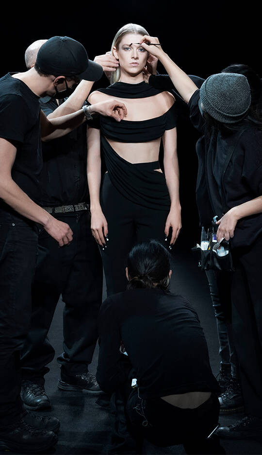 A model stands confidently while several stylists adjust her outfit and makeup in a dark studio setting.