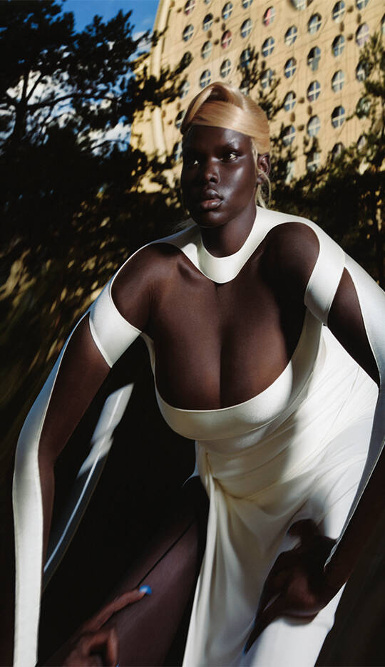 A model poses dramatically outdoors, wearing a flowing white dress with unique cutouts. The sun creates striking shadows, highlighting her features against a blurred urban backdrop.