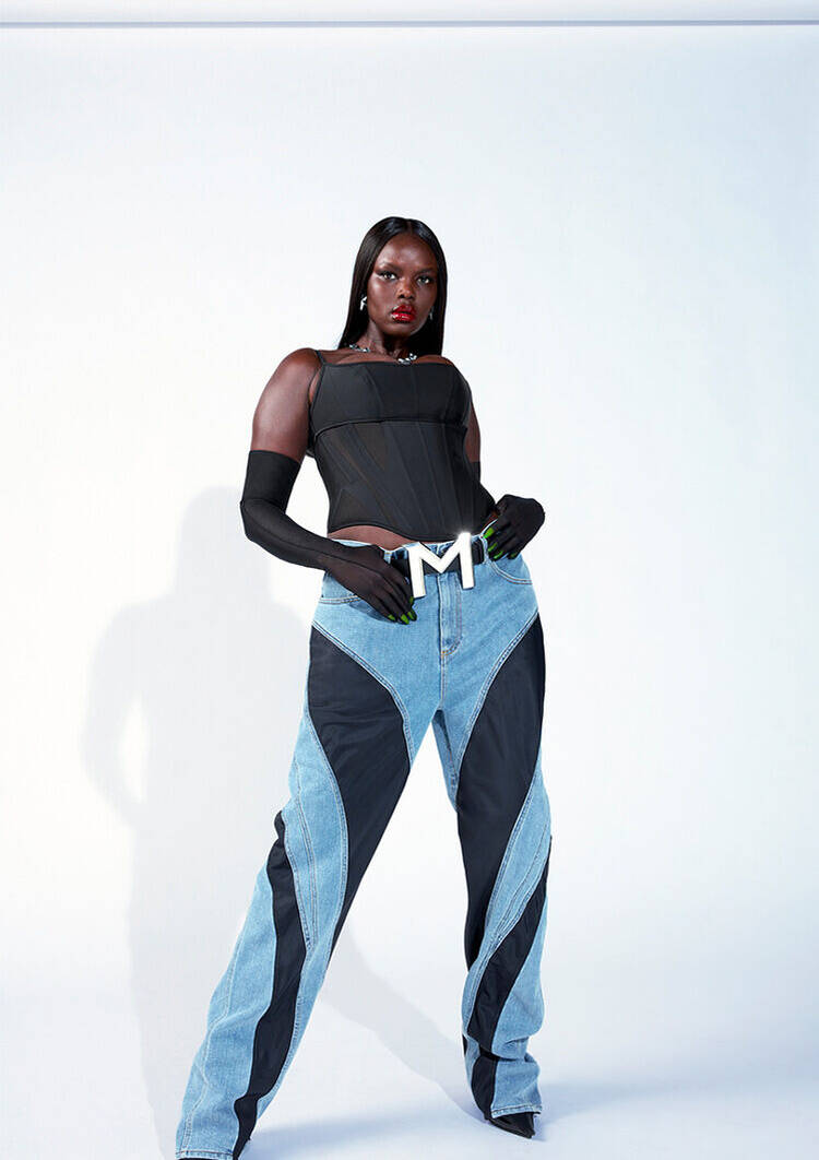 A model poses confidently against a light background, wearing a black corset top, black gloves, and oversized denim pants with a distinctive design. The outfit features a prominent belt buckle with the letter "M."