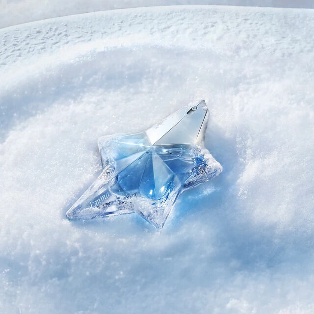 A close-up shot of an engraved iconic light blue, star-shaped Mugler Angel perfume bottle resting elegantly on a bed of pristine, sparkling white snow under soft blue light.
