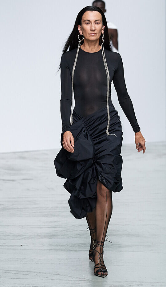 A model walks down a runway wearing a fitted black long-sleeve top and a dramatic, asymmetrical black skirt with ruffles, paired with strappy heels and statement earrings.