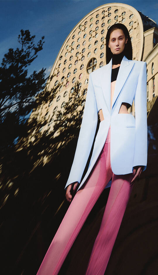 A model wearing a light blue blazer with cut-out details and pink pants stands against a backdrop of a uniquely shaped building. The image captures a blend of fashion and architecture, with a dramatic perspective.