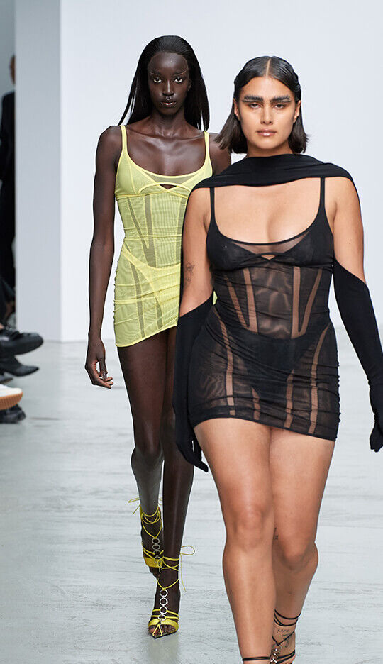 Two models walk down a runway during a fashion show. The first model, wearing a sheer yellow dress, leads; the second model follows in a black sheer dress with long sleeves. The setting features a minimalist backdrop.