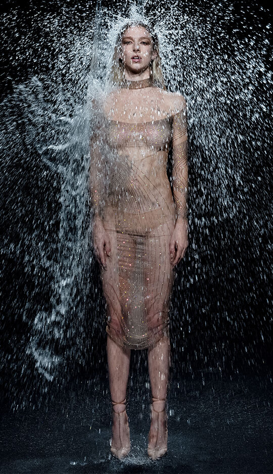 A model stands confidently in a sheer, sparkling outfit while water cascades around her, creating a dynamic and dramatic effect against a dark background.