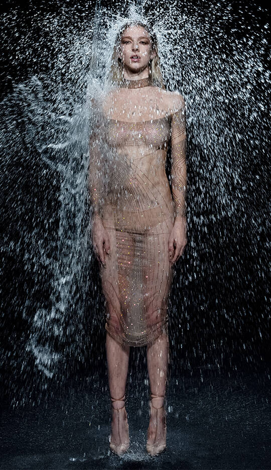 A model stands confidently in a sheer, sparkling outfit while water cascades around her, creating a dynamic and dramatic effect against a dark background.