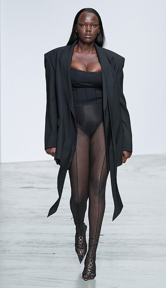 A model walks down the runway wearing a black oversized blazer over a fitted black bodysuit and sheer, high-waisted tights, paired with stylish heels. The setting is a minimalist fashion show with a plain backdrop.