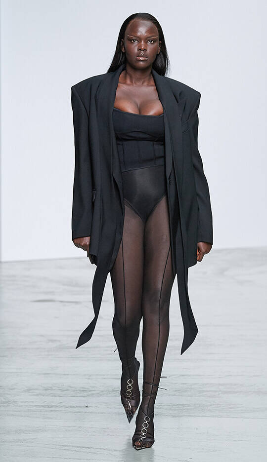 A model walks down the runway wearing a black oversized blazer over a fitted black bodysuit and sheer, high-waisted tights, paired with stylish heels. The setting is a minimalist fashion show with a plain backdrop.