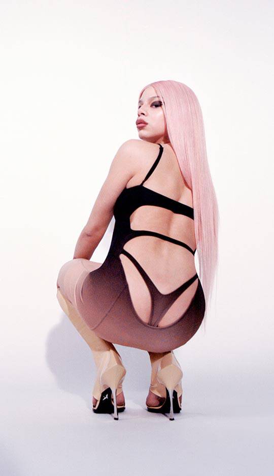 A person with long pink hair poses from the back, wearing a black and beige gradient outfit and high heels, highlighting a stylish and confident stance.