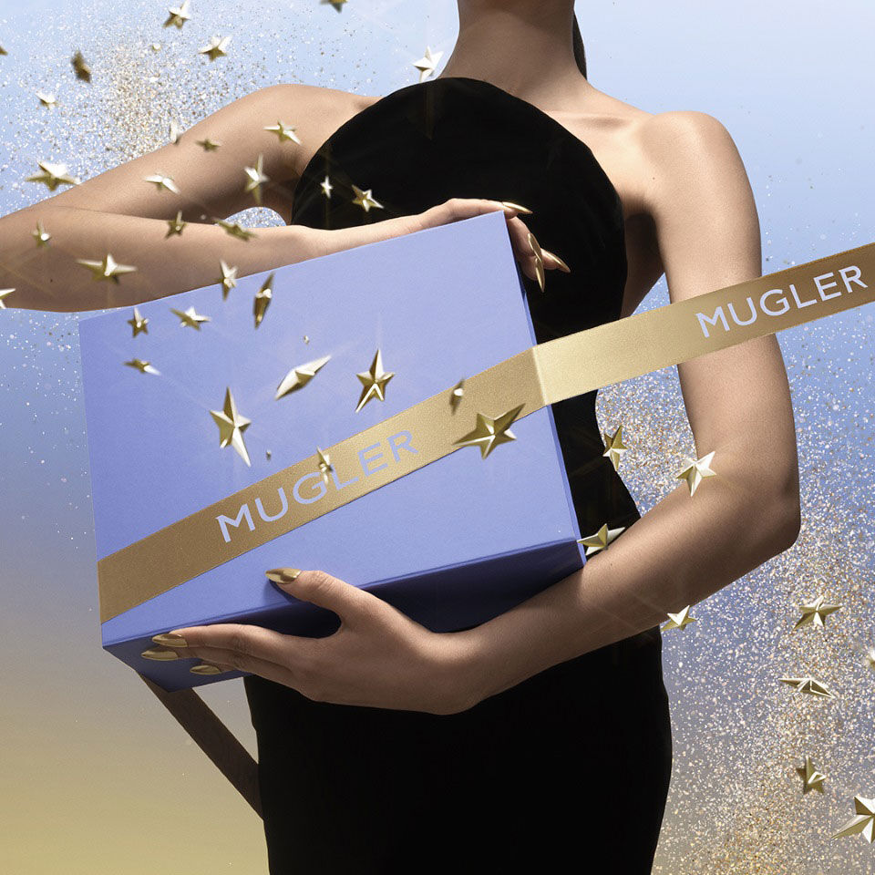 A person in a black dress cradles a light‑blue Mugler gift box wrapped with a gold Mugler ribbon, surrounded by floating gold stars and glitter against a blue‑to‑gold gradient sky.