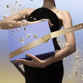 A person in a black dress cradles a light‑blue Mugler gift box wrapped with a gold Mugler ribbon, surrounded by floating gold stars and glitter against a blue‑to‑gold gradient sky.