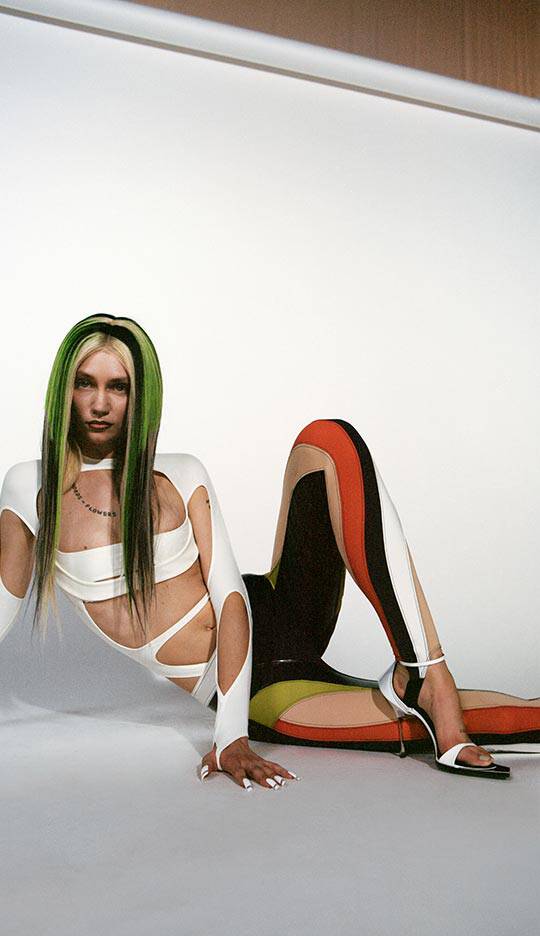 A model poses seated on the floor, wearing a stylish outfit with cutouts and contrasting colors, featuring long green hair and a confident expression against a plain background.