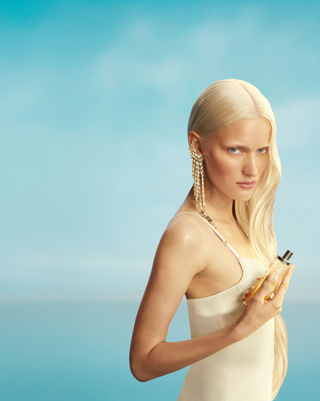 New Mugler Starlicious les eaux gourmandes campaign banner featuring a model with long blonde hair and blue eyes, wearing a pale yellow dress, holding the Pineapple Musk fragrance bottle against a soft blue sky background. The fragrances are delicious, collectable and layerable. 