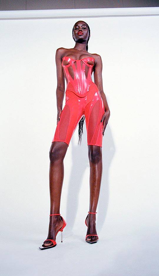 A tall model poses in a striking red latex outfit with a strapless design and thigh-high cut, showcasing sleek styling and high heels against a plain white background.