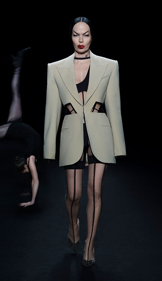 A model walks down the runway wearing a tailored cream blazer with cutouts, paired with a black mini dress and thigh-high stockings, while another figure performs a dramatic pose in the background. The scene is set against a dark backdrop, highlighting the striking fashion.
