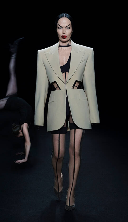 A model walks down the runway wearing a tailored cream blazer with cutouts, paired with a black mini dress and thigh-high stockings, while another figure performs a dramatic pose in the background. The scene is set against a dark backdrop, highlighting the striking fashion.