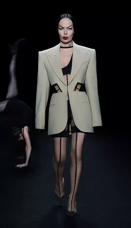 A model walks down the runway wearing a tailored cream blazer with cutouts, paired with a black mini dress and thigh-high stockings, while another figure performs a dramatic pose in the background. The scene is set against a dark backdrop, highlighting the striking fashion.