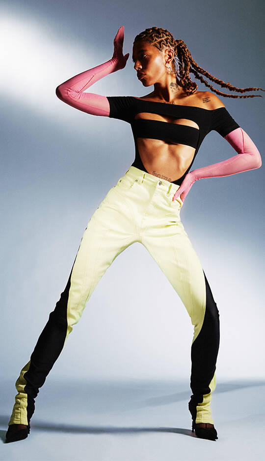 A model poses dramatically in a black and yellow outfit, featuring an off-the-shoulder top with cutouts and high-waisted, contrasting pants. She wears long pink gloves and striking heels, striking a bold pose against a softly lit background.
