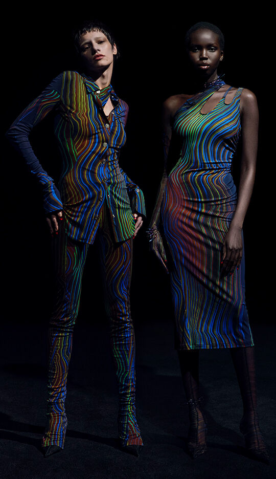 Two models stand against a dark background, wearing vibrant, patterned outfits that feature colorful, swirling designs. One model is dressed in a form-fitting shirt and pants, while the other wears a sleek, asymmetrical dress.