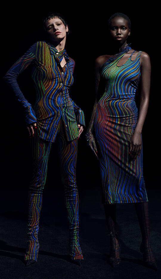 Two models stand against a dark background, wearing vibrant, patterned outfits that feature colorful, swirling designs. One model is dressed in a form-fitting shirt and pants, while the other wears a sleek, asymmetrical dress.