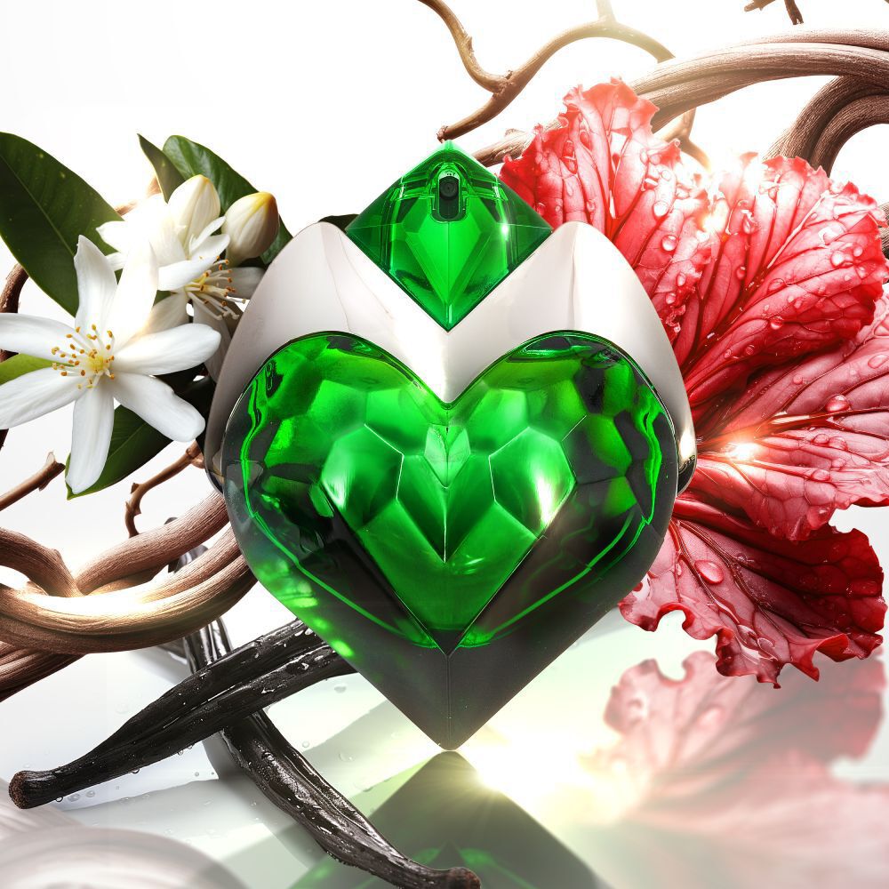 A heart-shaped green faceted perfume bottle with a white and green cap, surrounded by white jasmine flowers, a large red leaf, entwined wooden branches, and dark vanilla bean pods, set against a bright white background.