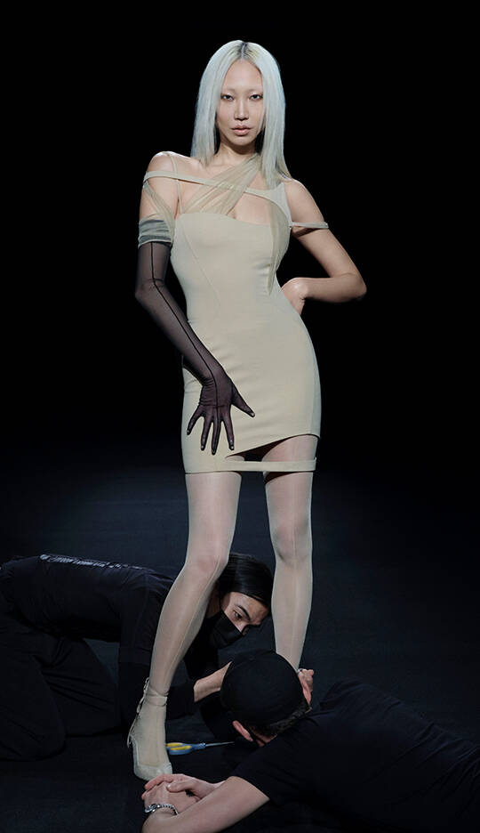 A model with long white hair poses confidently in a form-fitting beige dress, featuring sheer elements. Two individuals in black clothing are positioned at her feet, creating a dramatic scene against a dark background.