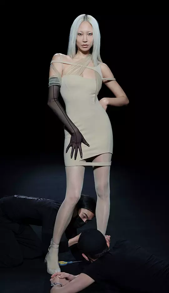 A model with long white hair poses confidently in a form-fitting beige dress, featuring sheer elements. Two individuals in black clothing are positioned at her feet, creating a dramatic scene against a dark background.