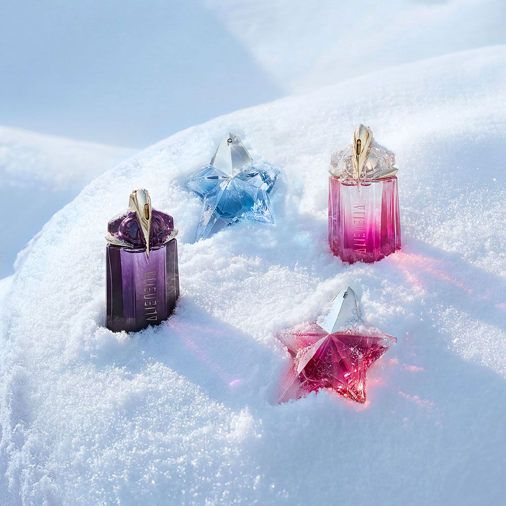 A purple Mugler Alien bottle, a light blue Angel bottle, a pink Alien bottle, and a pink Angel bottle beautifully spaced out on a soft, undulating snow surface. image redirects to our gift guide