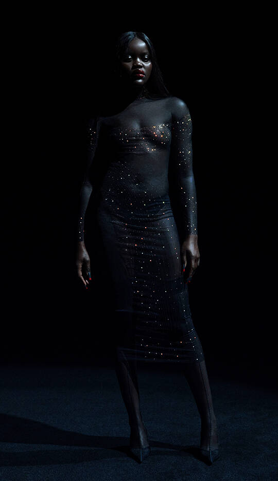 A model stands confidently in a sheer, embellished black dress against a dark background, emanating an elegant and striking presence.