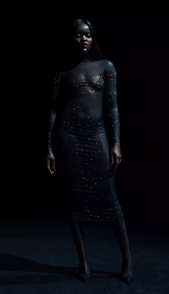 A model stands confidently in a sheer, embellished black dress against a dark background, emanating an elegant and striking presence.