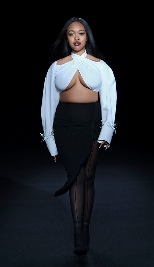 A model walks down a dark runway wearing a stylish outfit composed of a white top with unique cutouts and a black skirt, showcasing a modern and high-fashion aesthetic.