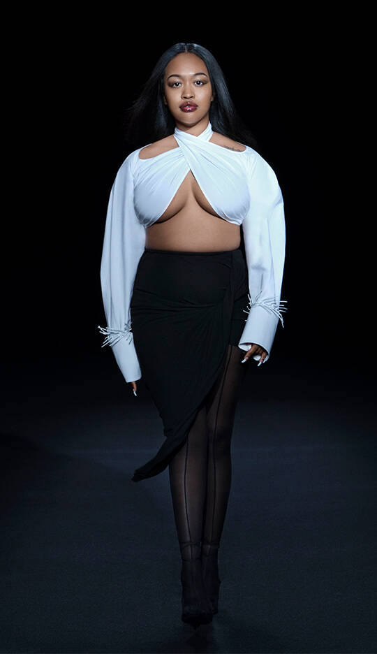 A model walks down a dark runway wearing a stylish outfit composed of a white top with unique cutouts and a black skirt, showcasing a modern and high-fashion aesthetic.
