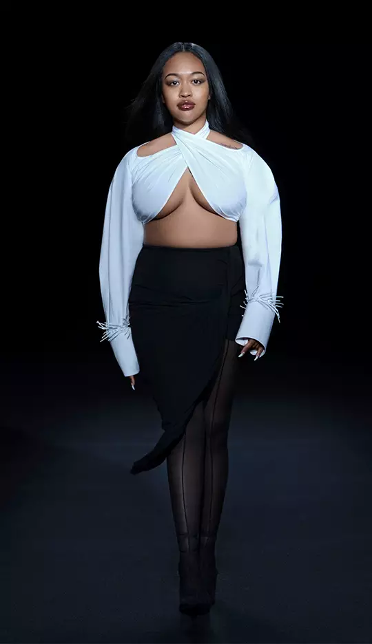A model walks down a dark runway wearing a stylish outfit composed of a white top with unique cutouts and a black skirt, showcasing a modern and high-fashion aesthetic.